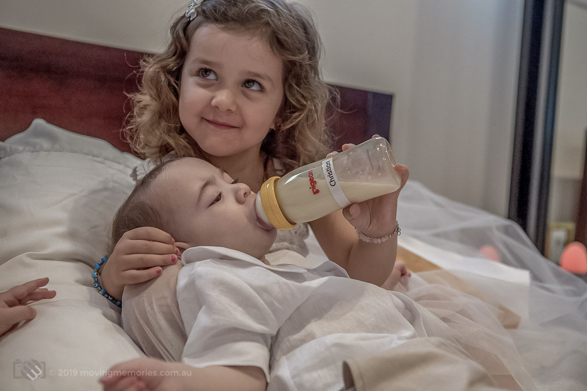 a twin baby boy Christian having some precious time and fed milk from a bottle by a young girl at home before his Greek Orthodox Christening in Sydney