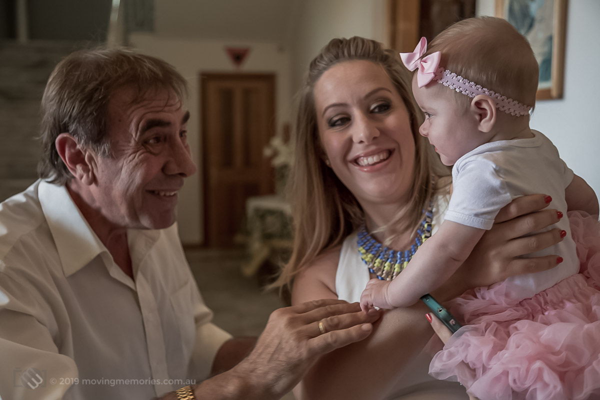 a baby girl held by her mother as the proud grand father gets a smile from the baby girl at a Greek Orthodox Church in Sydney