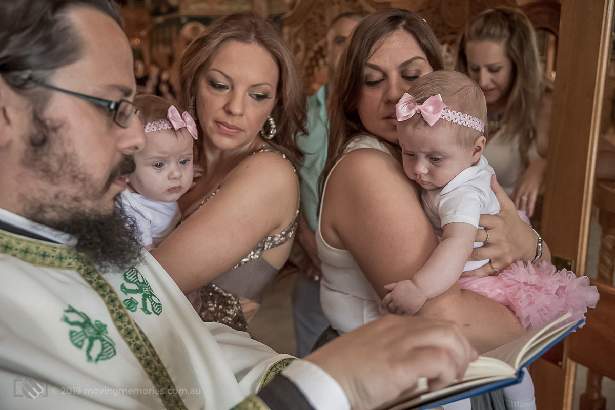 twin baby girls with their God mothers at the entrance to the Greek Orthodox Church with the priest pray and repeat affirmations at the start of the Greek Orthodox ceremony in Sydney
