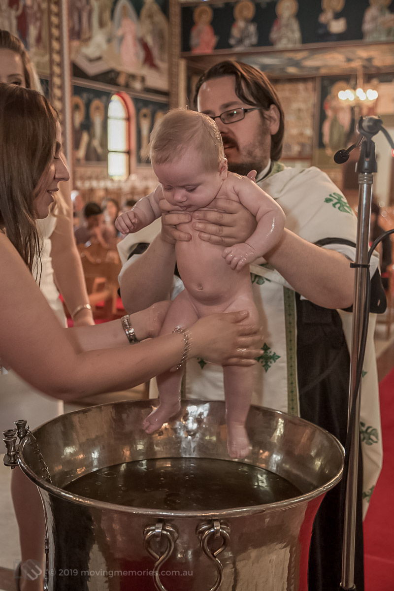 baby girl covered in oil by the God mother just before the baby is immersed in water at the baptism font Sydney Greek Orthodox Christening Moving Memories Australia